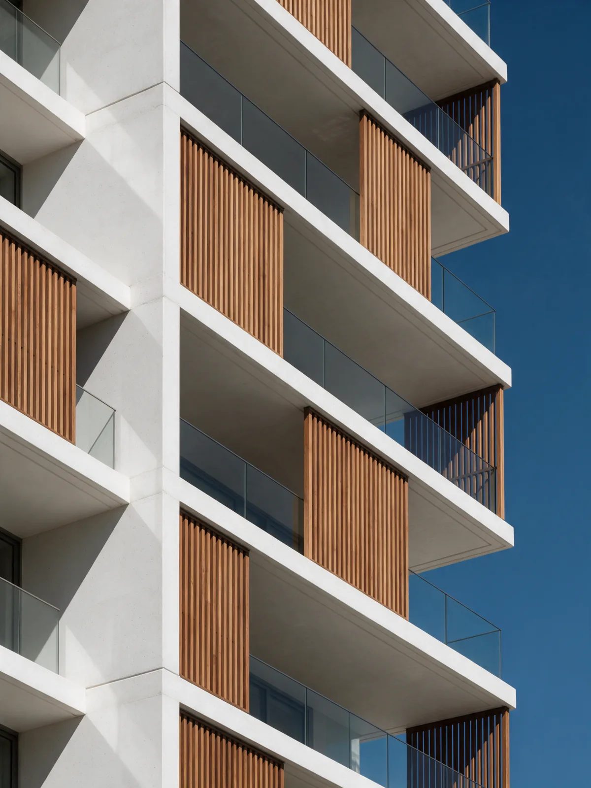 Detalle de la fachada: balcones de concreto blanco en voladizo, barandas de vidrio y celosías verticales de madera contra el cielo azul.