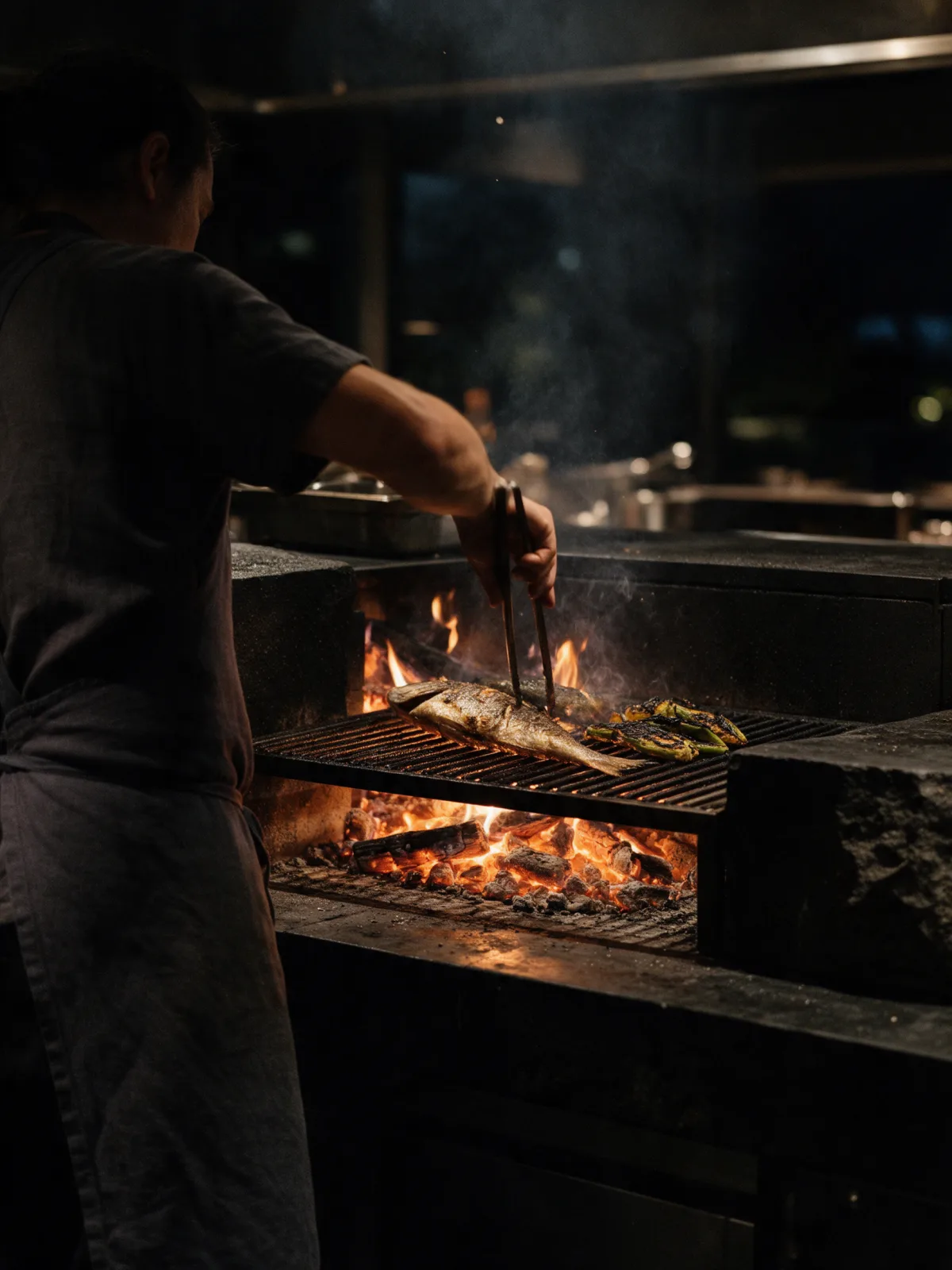 Cocinero de espaldas volteando un pescado entero con pinzas largas sobre la parrilla de leña, entre llamas, brasas y humo